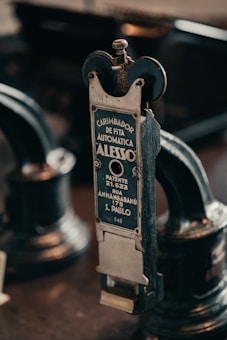 A vintage metal device with a label in Portuguese indicating it is an automatic ribbon stamper patented in São Paulo. The intricate machinery features wheels and knobs, emphasizing its industrial and historical design.
