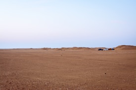 A vast desert landscape stretches out with a flat sandy foreground and soft, undulating dunes in the distance. To the right, two vehicles, one white and one dark, are parked near a small tent, suggesting a campsite. The sky above is clear with a soft gradient from blue to light pink.