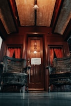 A vintage interior with dark wooden elements, featuring two wicker chairs flanking a wooden door. The space is decorated with red curtains and a sign on the door reading 'PROIBIDO ULTRAPASSAR A GRADE'. The ceiling has a warm, dim light.