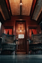 A vintage interior with dark wooden elements, featuring two wicker chairs flanking a wooden door. The space is decorated with red curtains and a sign on the door reading 'PROIBIDO ULTRAPASSAR A GRADE'. The ceiling has a warm, dim light.