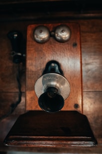 A vintage wall-mounted telephone features a wooden base with metallic bells and a protruding mouthpiece and earpiece. The design is reminiscent of early 20th-century communication devices, reflecting a sense of nostalgia and antique charm.