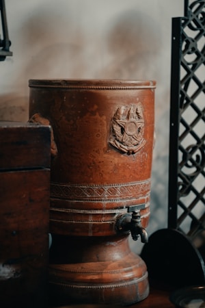 A rustic, vintage-style ceramic urn with ornate detailing and a prominent emblem sits on a wooden surface. It features a metal spigot at the base and is positioned against a wall with decorative metalwork nearby.