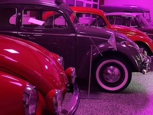 A row of vintage cars with a focus on a classic Volkswagen Beetle in the foreground. The image is illuminated with bright pink lighting, casting colorful reflections on the shiny surfaces of the cars. Each vehicle displays features characteristic of mid-20th century automobile design, such as rounded headlights and chrome accents.
