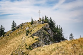 A cross stands atop a grassy hill surrounded by tall pine trees. The hill is rocky, with a narrow winding path leading up to the peak. A fence runs along the path. The sky is partly cloudy, creating a serene and peaceful atmosphere.