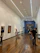A modern art gallery with wooden flooring and high ceiling, showcasing several paintings on the walls. There are a few people walking around and viewing the artwork. The lighting is bright, emphasizing the artwork.