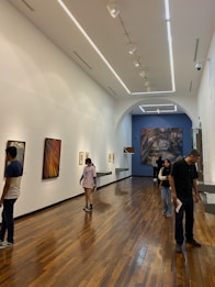 A modern art gallery with wooden flooring and high ceiling, showcasing several paintings on the walls. There are a few people walking around and viewing the artwork. The lighting is bright, emphasizing the artwork.