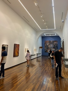 A modern art gallery with wooden flooring and high ceiling, showcasing several paintings on the walls. There are a few people walking around and viewing the artwork. The lighting is bright, emphasizing the artwork.