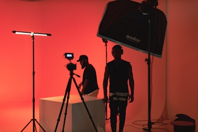 A dimly lit studio scene with red ambient lighting. Two people are present, one is standing near a camera setup, while the other is adjusting equipment on a table. Studio lighting equipment and softboxes are visible, contributing to the overall moody and artistic atmosphere.