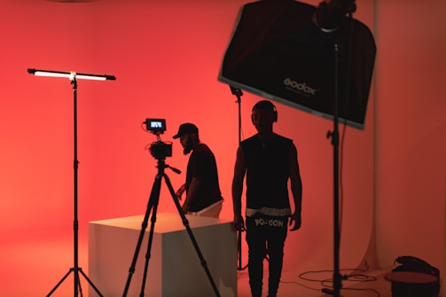 A dimly lit studio scene with red ambient lighting. Two people are present, one is standing near a camera setup, while the other is adjusting equipment on a table. Studio lighting equipment and softboxes are visible, contributing to the overall moody and artistic atmosphere.