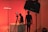 A dimly lit studio scene with red ambient lighting. Two people are present, one is standing near a camera setup, while the other is adjusting equipment on a table. Studio lighting equipment and softboxes are visible, contributing to the overall moody and artistic atmosphere.