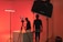 A dimly lit studio scene with red ambient lighting. Two people are present, one is standing near a camera setup, while the other is adjusting equipment on a table. Studio lighting equipment and softboxes are visible, contributing to the overall moody and artistic atmosphere.