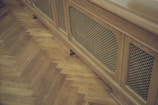 A wooden radiator cover with a lattice design is positioned against a parquet floor. The floor features a herringbone pattern, highlighting the warm tones of the wood.