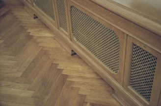 A wooden radiator cover with a lattice design is positioned against a parquet floor. The floor features a herringbone pattern, highlighting the warm tones of the wood.