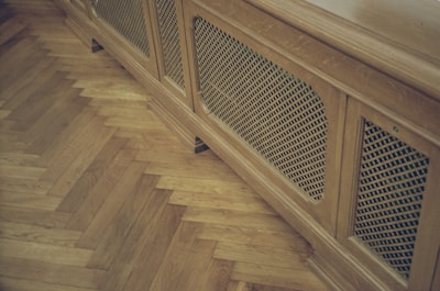 A wooden radiator cover with a lattice design is positioned against a parquet floor. The floor features a herringbone pattern, highlighting the warm tones of the wood.