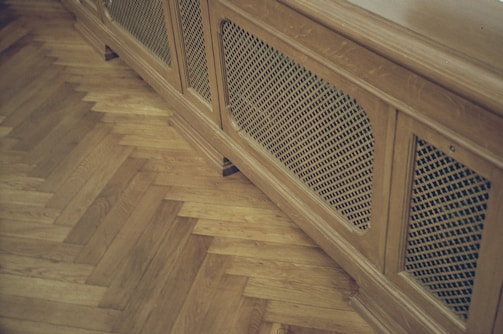 A wooden radiator cover with a lattice design is positioned against a parquet floor. The floor features a herringbone pattern, highlighting the warm tones of the wood.