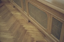 A wooden radiator cover with a lattice design is positioned against a parquet floor. The floor features a herringbone pattern, highlighting the warm tones of the wood.