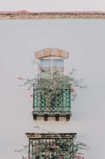 A green balcony adorned with lush climbing plants and pink flowers is attached to a white wall. Above the balcony, there is a window with a brick arch. The exterior design combines rustic and natural elements.