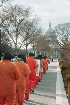 A group of people dressed in orange jumpsuits and black head coverings are walking in a line along a sidewalk. They are holding signs. Leafless trees line the pathway with a dome-like building in the distance.