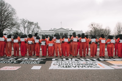 A group of individuals in orange jumpsuits and black hoods stand in a line holding signs in front of the White House. Large black and white banners on the ground advocate for closing Guantanamo and justice for its survivors. Leafless trees frame the scene against a cloudy sky.
