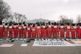 A group of individuals in orange jumpsuits and black hoods stand in a line holding signs in front of the White House. Large black and white banners on the ground advocate for closing Guantanamo and justice for its survivors. Leafless trees frame the scene against a cloudy sky.