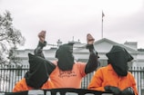 Three individuals wearing orange jumpsuits and black hoods stand in front of a large white building, likely a government structure. The central figure is raising their hands, which are shackled, and the shirt reads 'SHUT DOWN GUANTANAMO'. The scene suggests a protest or demonstration with a focus on human rights or political issues.