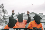 Three individuals wearing orange jumpsuits and black hoods stand in front of a large white building, likely a government structure. The central figure is raising their hands, which are shackled, and the shirt reads 'SHUT DOWN GUANTANAMO'. The scene suggests a protest or demonstration with a focus on human rights or political issues.