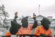 Three individuals wearing orange jumpsuits and black hoods stand in front of a large white building, likely a government structure. The central figure is raising their hands, which are shackled, and the shirt reads 'SHUT DOWN GUANTANAMO'. The scene suggests a protest or demonstration with a focus on human rights or political issues.