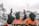 Three individuals wearing orange jumpsuits and black hoods stand in front of a large white building, likely a government structure. The central figure is raising their hands, which are shackled, and the shirt reads 'SHUT DOWN GUANTANAMO'. The scene suggests a protest or demonstration with a focus on human rights or political issues.