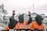 Three individuals wearing orange jumpsuits and black hoods stand in front of a large white building, likely a government structure. The central figure is raising their hands, which are shackled, and the shirt reads 'SHUT DOWN GUANTANAMO'. The scene suggests a protest or demonstration with a focus on human rights or political issues.