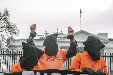 Three individuals wearing orange jumpsuits and black hoods stand in front of a large white building, likely a government structure. The central figure is raising their hands, which are shackled, and the shirt reads 'SHUT DOWN GUANTANAMO'. The scene suggests a protest or demonstration with a focus on human rights or political issues.