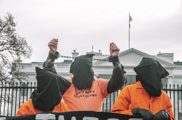 Three individuals wearing orange jumpsuits and black hoods stand in front of a large white building, likely a government structure. The central figure is raising their hands, which are shackled, and the shirt reads 'SHUT DOWN GUANTANAMO'. The scene suggests a protest or demonstration with a focus on human rights or political issues.