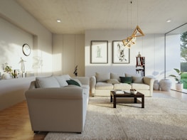 A modern living room with a minimalist aesthetic features light-colored sofas accented with green and beige pillows. The room includes a dark wooden coffee table on a textured area rug. Artwork adorns the walls above the sofa, and a geometric pendant light hangs from the ceiling. Large windows allow natural light to flood the space, and various potted plants enhance the decor. A bookshelf and decorative items are neatly arranged in the corner, and a wall clock is visible.