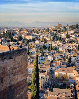 A picturesque cityscape featuring a densely packed collection of buildings with terracotta roofs, interspersed with tall cypress trees. The city is set against a backdrop of distant mountains under a clear blue sky. In the foreground, people are seen standing on an ancient stone wall enjoying the panoramic view.