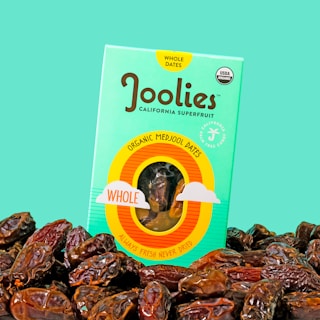 A package of organic Medjool dates labeled 'Joolies' is prominently displayed against a solid teal background. The bright packaging features a window that reveals the dates inside and is accented with text emphasizing that the dates are fresh and never dried. A pile of whole dates surrounds the package in the foreground.
