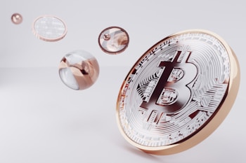A large, stylized Bitcoin symbol is depicted on a golden coin, surrounded by several floating, reflective spheres with metallic and transparent surfaces.
