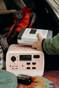 A person is placing a Newpowa solar charge controller on top of a portable power station inside a vehicle. There is a colorful patterned blanket beneath the power station and a stainless steel water bottle with stickers nearby. A red and gray backpack is in the background.