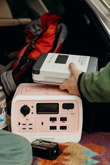 A person is placing a Newpowa solar charge controller on top of a portable power station inside a vehicle. There is a colorful patterned blanket beneath the power station and a stainless steel water bottle with stickers nearby. A red and gray backpack is in the background.