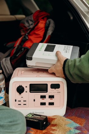 A person is placing a Newpowa solar charge controller on top of a portable power station inside a vehicle. There is a colorful patterned blanket beneath the power station and a stainless steel water bottle with stickers nearby. A red and gray backpack is in the background.