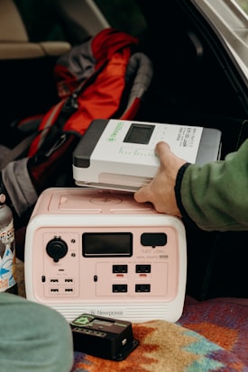 A person is placing a Newpowa solar charge controller on top of a portable power station inside a vehicle. There is a colorful patterned blanket beneath the power station and a stainless steel water bottle with stickers nearby. A red and gray backpack is in the background.