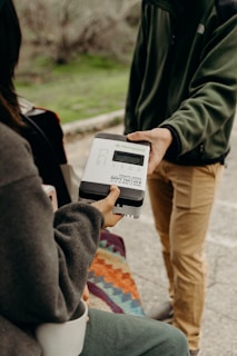 Two individuals are exchanging a device outdoors, one person holding a box-like gadget with labels, while the other reaches out to take it. A colorful patterned cloth can be seen in the foreground, and the background is blurred greenery.