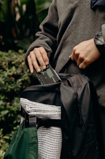 A person wearing a dark green fleece holds a handheld device near an open black backpack with a patterned pouch. The scene is set outdoors, with green foliage in the background.