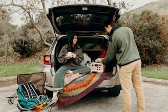 A person sits in the back of an SUV holding a mug, while another person stands next to the car arranging items. The open trunk is lined with a colorful blanket, and camping gear is visible including a backpack and folded chair. The scene takes place in an outdoor, wooded area.