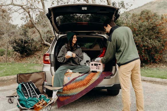 A person sits in the back of an SUV holding a mug, while another person stands next to the car arranging items. The open trunk is lined with a colorful blanket, and camping gear is visible including a backpack and folded chair. The scene takes place in an outdoor, wooded area.