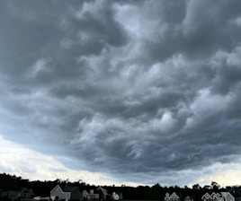 Dark, stormy clouds dominate the sky, swirling and creating a dramatic atmosphere. Below, a line of suburban houses with pitched roofs are lined up against a backdrop of trees. The scene suggests an impending storm, with the clouds appearing heavy and ominous.