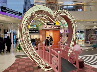 A heart-shaped, ornate archway illuminated by lights stands in the center of a busy shopping mall. Within the archway is a pink bridge leading to a photo booth decorated with cartoon characters and a welcome sign. Shoppers are visible in the background, moving through the brightly lit environment.