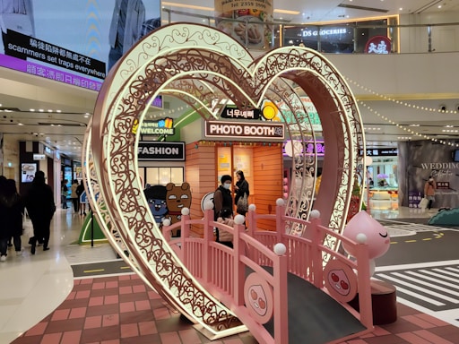 A heart-shaped, ornate archway illuminated by lights stands in the center of a busy shopping mall. Within the archway is a pink bridge leading to a photo booth decorated with cartoon characters and a welcome sign. Shoppers are visible in the background, moving through the brightly lit environment.