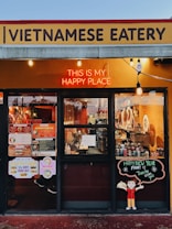 A Vietnamese eatery storefront features a sign with bold lettering. A neon sign reads 'This is my happy place' above large windows displaying the menu and festive decorations. The interior shows various items, including bottles and kitchen equipment. There is a cheerful cartoon character holding a dish on a board that wishes a happy new year.