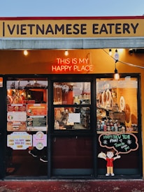 A Vietnamese eatery storefront features a sign with bold lettering. A neon sign reads 'This is my happy place' above large windows displaying the menu and festive decorations. The interior shows various items, including bottles and kitchen equipment. There is a cheerful cartoon character holding a dish on a board that wishes a happy new year.