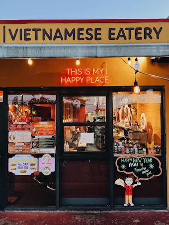 A Vietnamese eatery storefront features a sign with bold lettering. A neon sign reads 'This is my happy place' above large windows displaying the menu and festive decorations. The interior shows various items, including bottles and kitchen equipment. There is a cheerful cartoon character holding a dish on a board that wishes a happy new year.