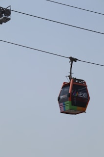 A colorful cable car hangs from a wire against a clear sky. The car features bright, vibrant stripes at the bottom and the words 'EDOMEX'. The sky is light blue, providing a minimalistic backdrop to the modern transportation method.